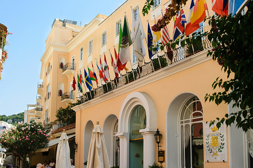 Capri, Italy - September 10, 2008: Hotel Located in Yellow Stucco Building, Capri, Italy. Capri is an island in the Tyrrhenian Sea next to Sorrentine Peninsula, in the Gulf of Naples in the Campania region of Italy. The main town of the island is Capri - which bears the same name as the island. It has been a resort since the time of the Roman Republic. Green trees, flags and clear blue clear sky are in the image.