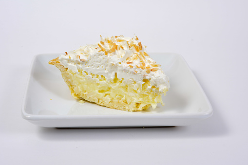 A slice of coconut cream pie with whipped cream topped with toated coconut shavings on a square white dessert plate