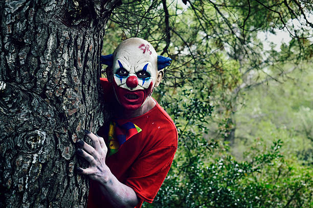 scary evil clown in the woods closeup of a scary evil clown in the woods, emerging from behind of a big tree face paint halloween adult men stock pictures, royalty-free photos & images