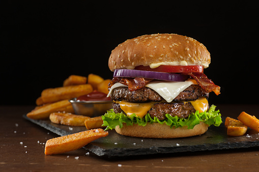 High resolution digital capture of a steakhouse-style double bacon cheeseburger with steak fries. This cheeseburger is made with two patties of ground steak, Cheddar and Monterey Jack cheeses, crispy bacon slices, fresh tomatoes, lettuce, and onion, all on a sesame seed bun. A ramekin of ketchup is visible amongst the fries. Background is dark and atmospheric, with room to expand and place copy.