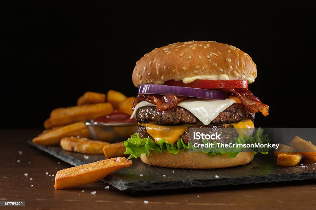 Steakhouse Double Bacon Cheeseburger High resolution digital capture of a steakhouse-style double bacon cheeseburger with steak fries. This cheeseburger is made with two patties of ground steak, Cheddar and Monterey Jack cheeses, crispy bacon slices, fresh tomatoes, lettuce, and onion, all on a sesame seed bun. A ramekin of ketchup is visible amongst the fries. Background is dark and atmospheric, with room to expand and place copy. Burger Stock Photo