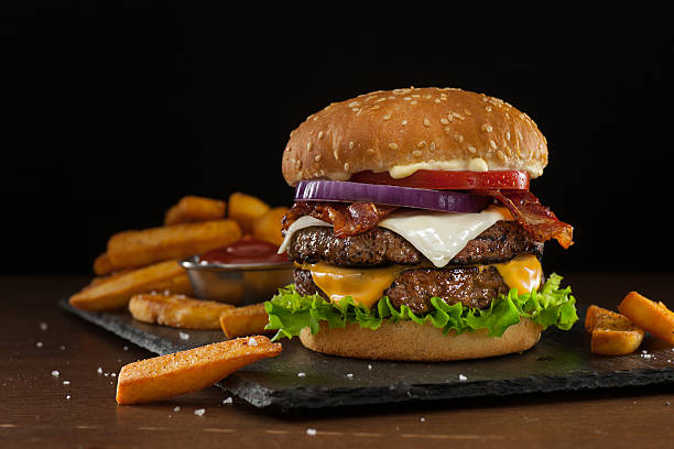 Steakhouse Double Bacon Cheeseburger High resolution digital capture of a steakhouse-style double bacon cheeseburger with steak fries. This cheeseburger is made with two patties of ground steak, Cheddar and Monterey Jack cheeses, crispy bacon slices, fresh tomatoes, lettuce, and onion, all on a sesame seed bun. A ramekin of ketchup is visible amongst the fries. Background is dark and atmospheric, with room to expand and place copy. cheddar cheese photos stock pictures, royalty-free photos & images