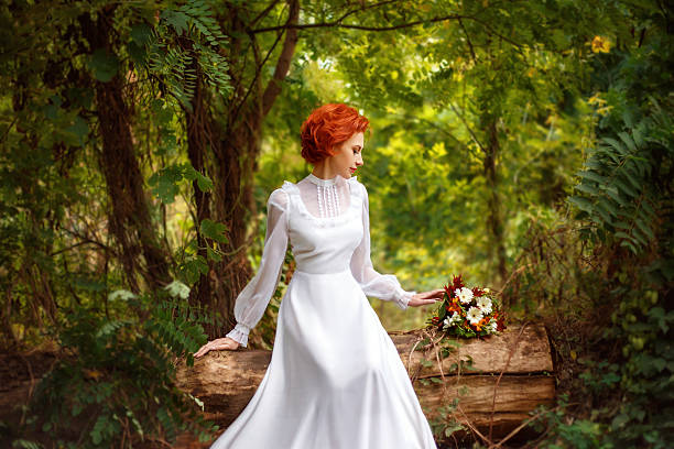 Gorgeous redheaded girl in white long dress sitting under the open sky and looking at the flowers. The picturesque nature. Fantastic shooting. Retro style. Fashionable toning. Creative color.