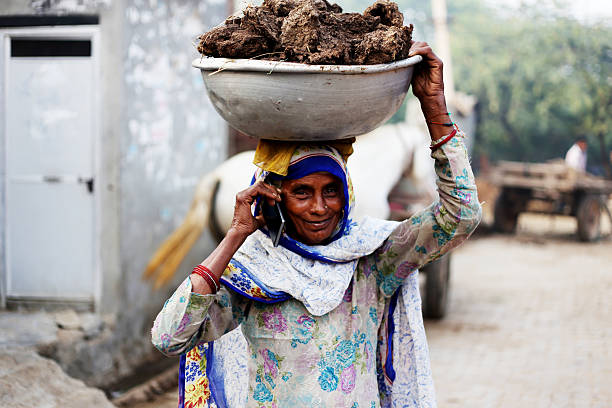 Old women carrying cow dung and talking on phone Rural women of India ethnicity wearing suit salwar and carrying buffalo dunk cakes on her head and talking on smart phone. cow dung stock pictures, royalty-free photos & images