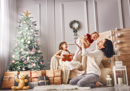 Merry Christmas and Happy Holidays! Cheerful mom and her cute daughters girls exchanging gifts. Parent and two little children having fun and playing together near Christmas tree indoors. Loving family with presents in room.