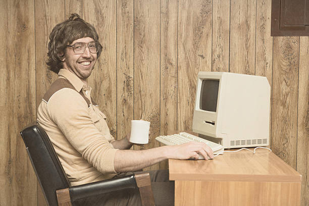 Retro Computer office nerd at home office A vintage retro styled image with brown tones of a man in a wood paneled 1980s room sitting at his computer desk with a coffee cup in hand smiling uncool stock pictures, royalty-free photos & images