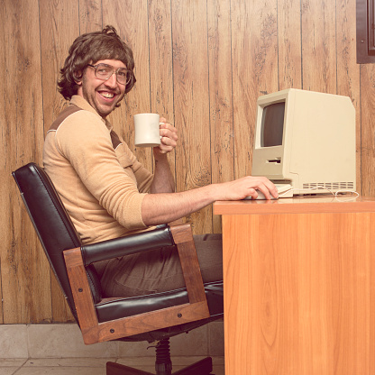 A vintage retro styled image with brown tones of a man in a wood paneled 1980s room sitting at his computer desk with a coffee cup in hand smiling