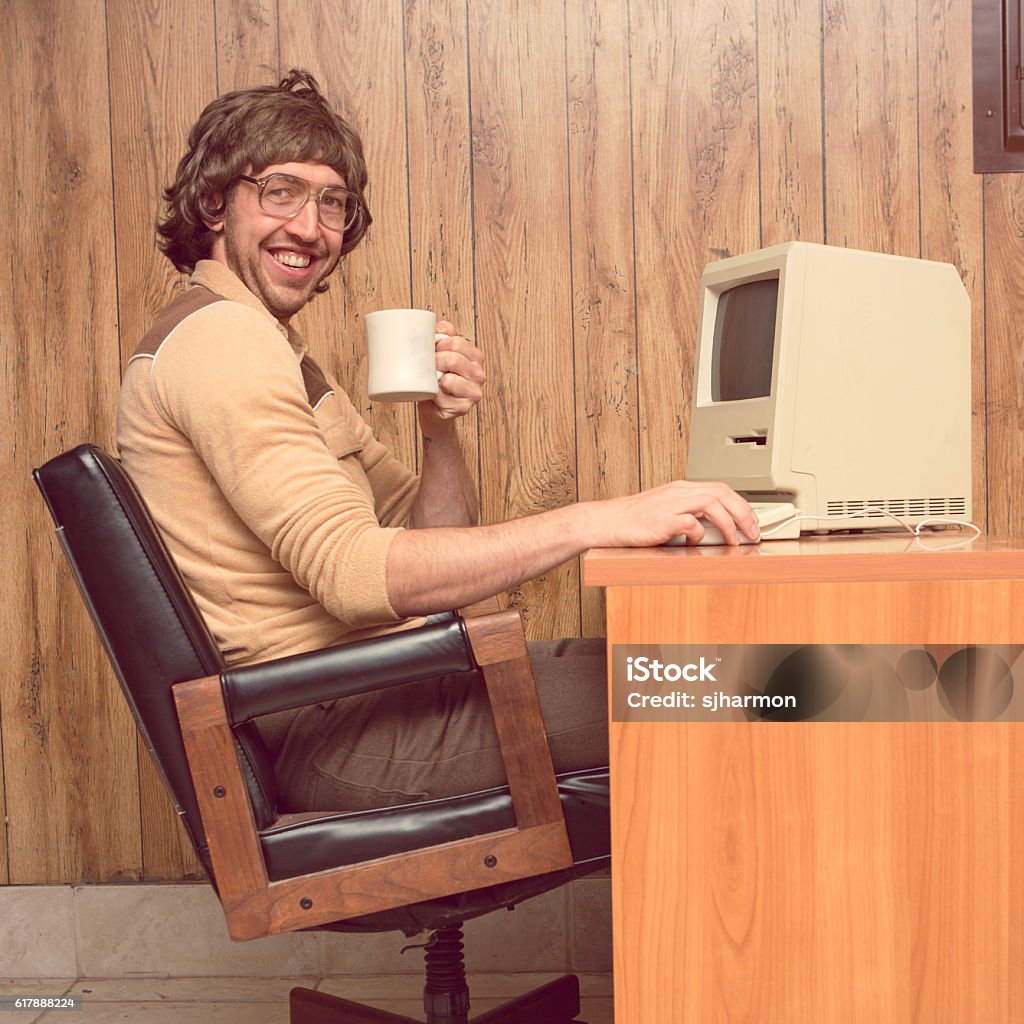 Funny 1980s Computer man at desk with coffee A vintage retro styled image with brown tones of a man in a wood paneled 1980s room sitting at his computer desk with a coffee cup in hand smiling Retro Style Stock Photo