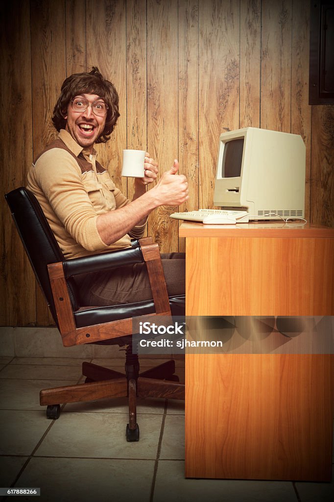 Exited Computer dorky nerd worker at desk A vintage retro styled image with brown tones of a man in a wood paneled 1980s room sitting at his computer desk with a coffee cup in hand smiling Humor Stock Photo