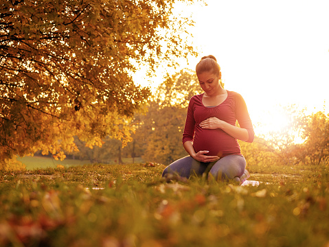Image of young beautiful woman in 30s, five or six months pregnant, walking in park and relaxing on beautiful autumn day. Woman is also practicing breathing exercises, yoga and meditation