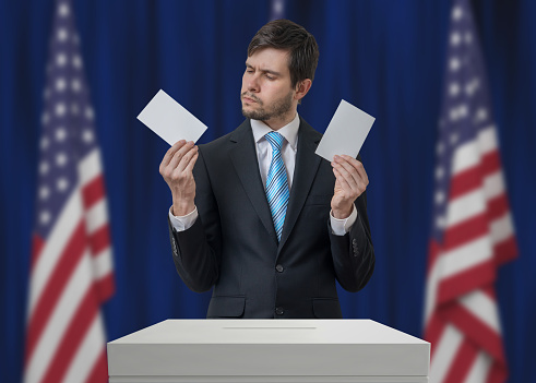 Election in United States of America. Undecided voter holds envelopes in hands above vote ballot and making decision. USA flags in background. Democracy concept.