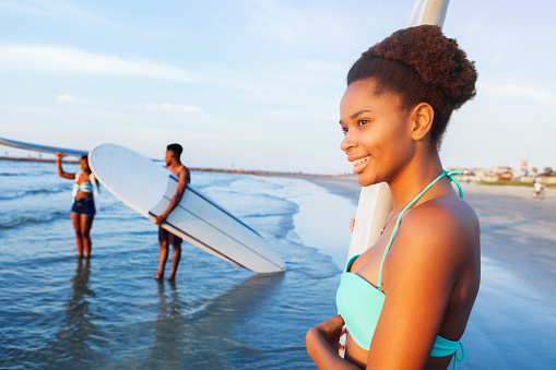 Cheerful African American teenage girl prepares to surf in the ocean with her friends. They are standing in shallow water holding surfboards. The girl is wearing a light green bikini. Her friends are talking in the background.