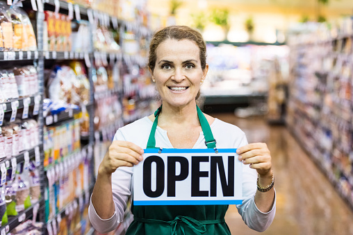 Smiling mature caucasian woman holding an open sign in a supermarket aisle