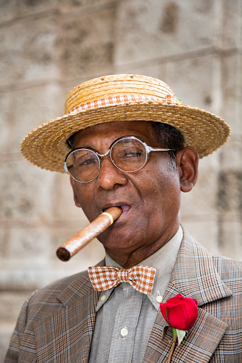 Portrait of an elegant, dark elderly Cuban gentleman in a suit, straw boater hat, and a cigar standing in front of a wall in Havana, Cuba