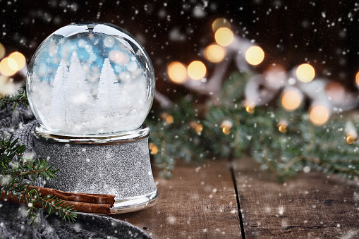 Rustic image of a snow globe surrounded by pine branches, cinnamon sticks and a warm gray scarf with falling snow. Shallow depth of field with selective focus on snowglobe.