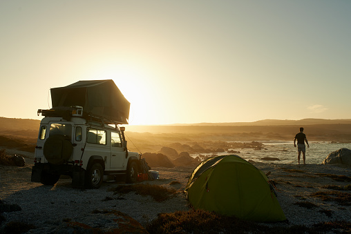 Shot of a silhouetted man admiring the view from his campsite by the sea