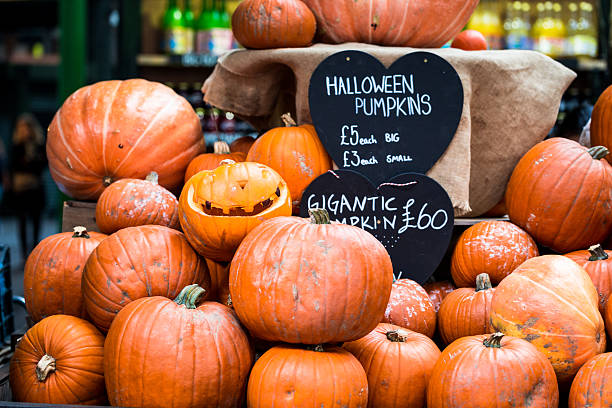 A display of large, small and carved pumpkins for Halloween in Borough Market, a famous food market in London, UK. There is a board showing the prices for the pumpkins, £3 for a small one, £5 for a big one, and £60 for a 'gigantic' pumpkin. Horizontal colour image with copy space.
