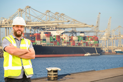 Dockworker standing in front of a container terminal in port. The man is wearing protective workwear and looking into the camera. A large container ship is loaded with cargo containers in the background while a fuel barge is refueling the ship. Overhead gantry cranes are loading and unloading the ships.