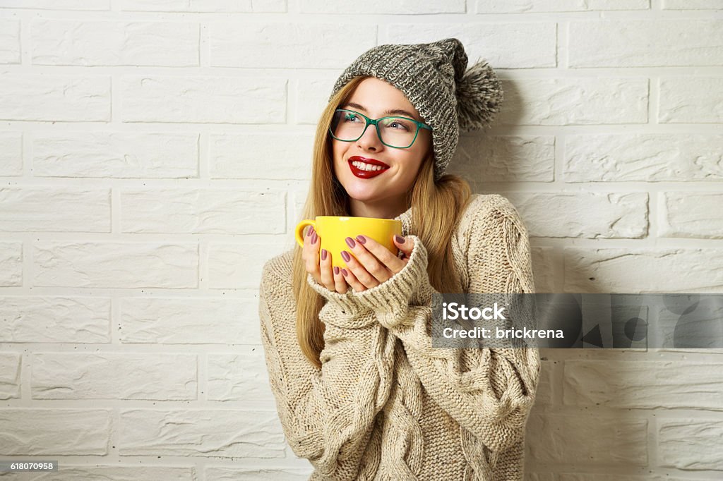 Smiling Hipster Girl in Winter Clothes with a Mug Happy Smiling Hipster Girl in Knitted Sweater and Beanie Hat with a Mug in Hands. Beautiful Woman at White Brick Wall Background. Winter or Autumn Warming Up Concept. Toned Photo with Copy Space. Eyeglasses Stock Photo