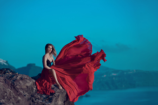 blond hair woman with red dress flying in the wind, looking away and posing on rock near the sea side.photo taken in Santorini.