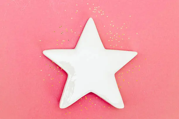 White ceramic star on pink background, with golden star-shaped glitter. Space for text.