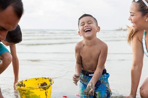Cute Hispanic boy smiles and laughs while playing in the sand with his family.
