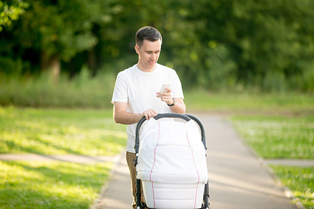Young father, dressed casually walking in park with baby carriage and holding mobile in hand, looking at screen smiling, messaging, using app