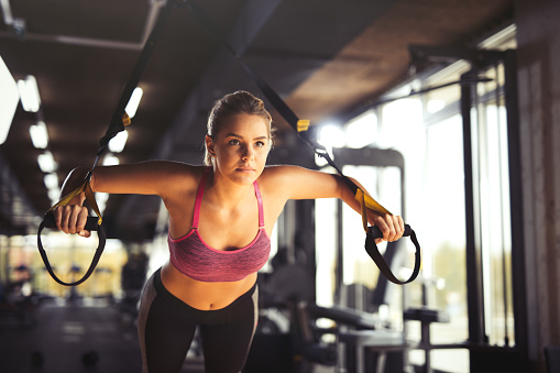 Young athletic woman having sports training and doing arm exercises with suspension straps exercises at gym.