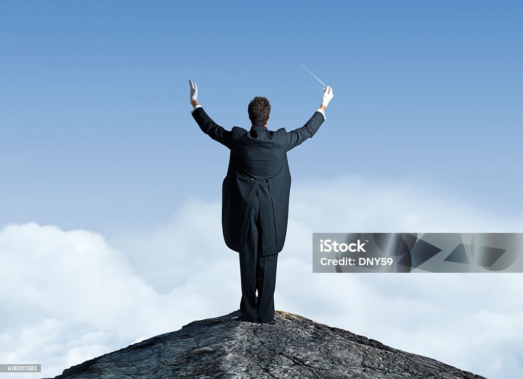 Musical Conductor On Top Of Mountain Top With Arms Raised A rear view of a musical conductor standing on top of a mountain top.  He has his arms raised with a baton in one hand as he is preparing to lead the orchestra.  Before him, and in the distance, white puffy clouds sit below a clear blue sky. Musical Conductor Stock Photo