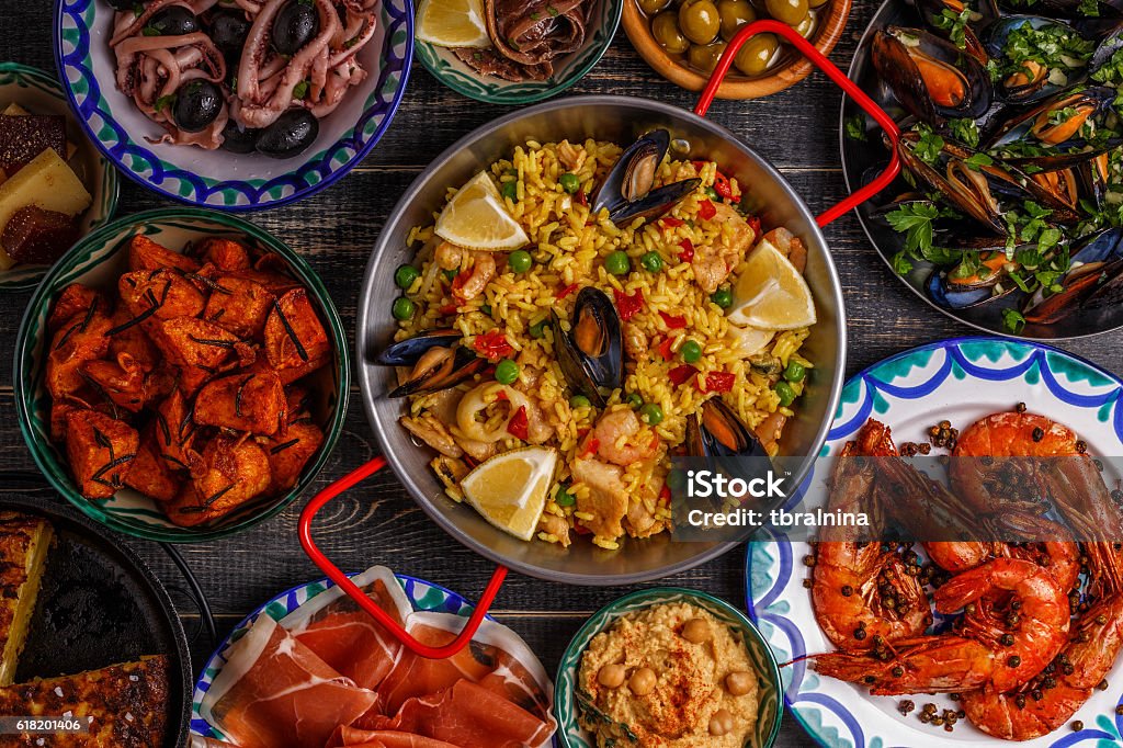 Typical spanish tapas concept, top view. Typical spanish tapas concept. Concept include slices jamon, bowls with olives,  anchovies, spicy potatoes, mashed chickpeas, shrimp, calamari, manchego with quince marmalade, pans with tortilla, paella, mussels  on a wooden table. Spain Stock Photo
