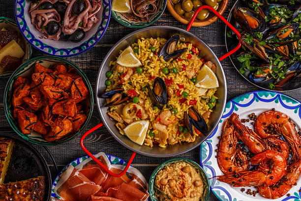 Typical spanish tapas concept, top view. Typical spanish tapas concept. Concept include slices jamon, bowls with olives,  anchovies, spicy potatoes, mashed chickpeas, shrimp, calamari, manchego with quince marmalade, pans with tortilla, paella, mussels  on a wooden table. spanish culture stock pictures, royalty-free photos & images