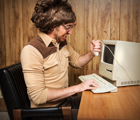 A retro looking funny angry computer office worker holding mouse and yelling in frustration.  Wood paneling office with vintage computer and keyboard on desk.
