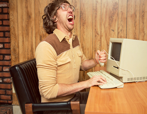 A retro looking funny angry computer office worker holding mouse and yelling in frustration.  Wood paneling office with vintage computer and keyboard on desk.