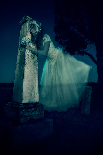 Female ghost bride in 20s dressed in a vintage wedding gown hovers and hugs an ancient grave  stone in a corn field in rural Indiana at night in an eerie greenish-blue light