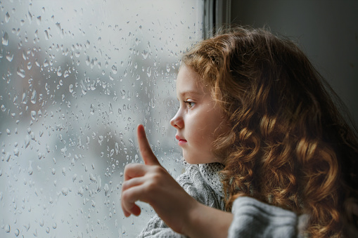 Sad little girl looking out the window on rain drops through wet glass autumn bad weather.