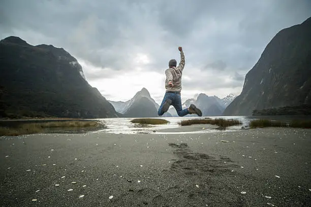 Cheering young man jumps in front of the Mitre peak in Milford sound, New Zealand.