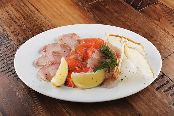 Smoked trout, grayling and whitefish with lemon slices on a white plate. Wooden background.