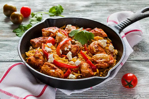 One-pot chicken fillet and orzo pasta with red bell peppers and feta cheese, cooked with garlic, paprika and olive oil. Cast-iron skillet and fresh tomatoes on wooden table.