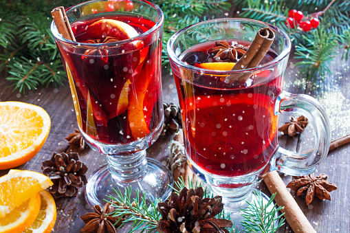 hot christmas mulled wine with snow, cinnamon sticks, anise and orange on wooden table background with fir branches and pine cones