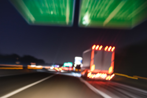 Defocused blurred motion of semi truck speeding on highway under street signs - Night traffic and transport logistic concept with semitruck container driving on speedway - Bokeh and tilted composition