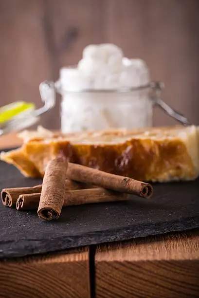 Vertical photo of apple strudel pie on black slate stone and wooden board. Few pieces of cinnamon next to sweets. White whipped cream in glass gar with open cover.