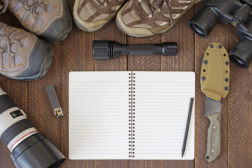 Top view of blank notebook with dirty hiking shoes, Flashlight, knife, tele lens, binocular, matchstick. Set of travel accessories flatlay on wooden background