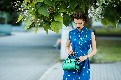 Attractive young woman in retro style blue dress with a green bag walking around the town on a sunny summer day. Portreit with red lips, retro hair, fashion look.