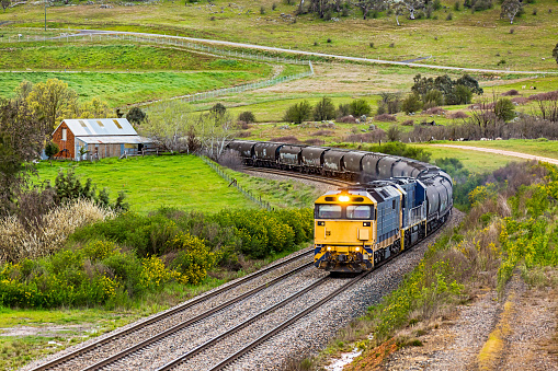Two powerful diesel locomotives haul a loaded grain train round curves and past old farm buildings and an overgrown caravan in lush green farmland on a wet, wintry spring day.  Horizontal, copy space.