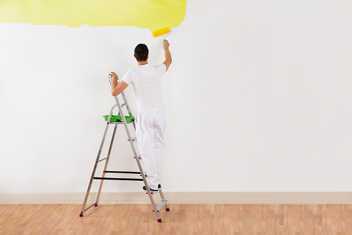Rear view of young man painting wall with yellow paint roller at home