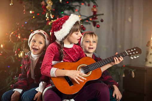 Little boys and little girl wearing santa's hat singing carols near christmas tree. The boys are aged 5 and the girl is aged 8. The girls is playing a guitar.