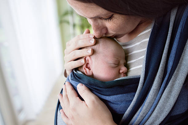 Close up, beautiful mother kissing her son in sling Close up, beautiful young mother kissing her newborn baby son in sling at home baby carrier stock pictures, royalty-free photos & images