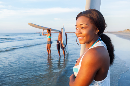 Confident teenage girl carries a white surfboard on the beach. She is wearing a white cover up over her swimsuit. Her friends are carrying their surfboards in the background.