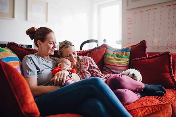 Same sex couple enjoying a cuddle together at home, with their baby daughter and their pet dog.