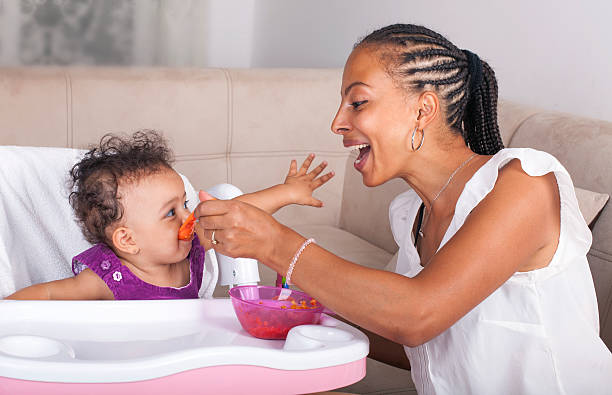 Mother Spoon Feeding Her Baby African-American Mother Feeding Baby Girl In High Chair high chair stock pictures, royalty-free photos & images